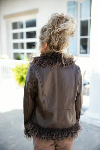 Back view of a woman wearing a dark brown faux leather jacket featuring prominent panel stitching across the back and long sleeves. Highlights the dramatic shaggy faux fur lining along the high collar stand and bottom hem, layered over brown denim pants.