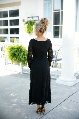 Back view of a woman wearing a long-sleeve black lace maxi dress, showcasing the full-coverage scooped back, back center seam zip closure, and semi-sheer lace sleeves. Styled with leopard print stiletto pumps and gold hoop earrings on a sunny outdoor patio walkway.