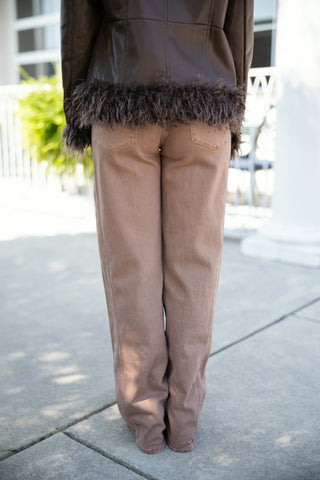 Back view focusing on high-waisted taupe-brown wide-leg denim jeans with classic back patch pockets, layered beneath a dark brown faux leather jacket trimmed with plush, shaggy faux fur along the lower hemline.