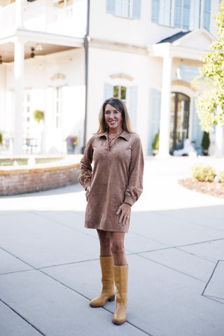 Full-length view of a woman modeling a taupe faux-suede long-sleeve shift dress resting above the knee. Styled with one hand in her pocket and paired with structured, light tan leather knee-high boots against an outdoor paved backdrop.