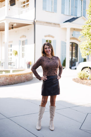 Woman posing outdoors in a brown long-sleeve snakeskin print top tucked into a brown faux-leather A-line mini skirt. She stands with hands on her hips wearing knee-high beige suede boots, layered gold necklaces with an oval pendant, and statement rings against a bright, modern architectural background.