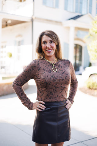 Close-up front view featuring a woman in a brown snakeskin printed long-sleeve crewneck top tucked into a structured brown leather skirt. Her hand rests on her hip, showcasing a stack of gold chain necklaces with a large oval pendant and decorative rings.