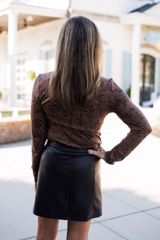 Back view of a woman wearing a brown snakeskin print long-sleeve top tucked into a brown leather mini skirt. One hand rests on her waist, showcasing the back seam line of the skirt and the fitted silhouette of the top.