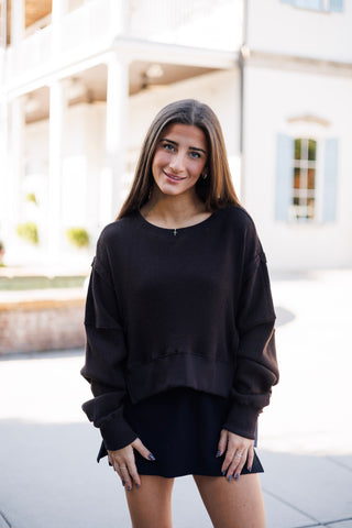 Woman smiling outdoors in a dark chocolate brown waffle-knit pullover top featuring drop shoulders, a crew neck, and raw exposed-seam detailing across the sleeves and hemline. Styled casually over a dark mini skirt or skort with a delicate cross pendant necklace in front of an elegant white building.