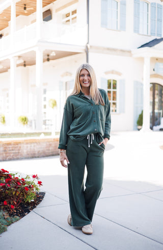 Woman posing outdoors in a dark forest green matching fleece lounge set, featuring a long-sleeve button-up shacket top and relaxed wide-leg pants with a white contrast drawstring waist. She stands with one hand in her pocket, wearing neutral beige sneakers in front of an elegant white architectural courtyard background.