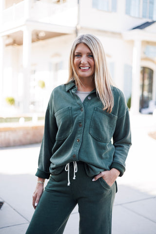 Frontal shot of a woman modeling a forest green fleece button-up lounge top layered over a neutral undershirt and paired with matching fleece wide-leg lounge trousers. Designed with dual chest patch pockets and a front-tuck styling with white drawstring waist details.