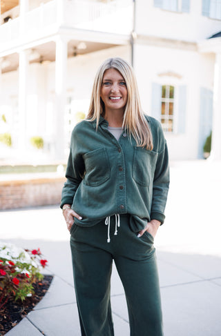 Frontal shot of a woman modeling a forest green fleece button-up lounge top layered over a neutral undershirt and paired with matching fleece wide-leg lounge trousers. Designed with dual chest patch pockets and a front-tuck styling with white drawstring waist details.