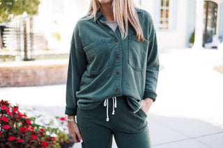 Detailed portrait highlighting the collar, button-front closure, and dual chest pockets of a dark green fleece shacket. Styled with rolled cuffs, a neutral crewneck base layer, and matching drawstring lounge pants.