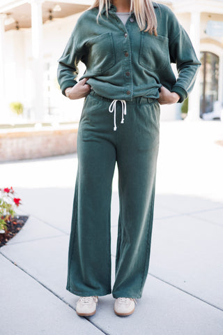Cropped front view emphasizing the high-waisted wide-leg fleece pants in dark forest green with a functional white drawstring cord. The matching button-up shacket is styled tucked at the waistband with hands resting near the pockets, paired with neutral beige low-top sneakers.