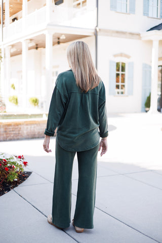 Back view of a woman wearing a dark forest green two-piece fleece set. The long-sleeve shirt top features a dropped shoulder design and a curved high-low hemline, paired with full-length wide-leg fleece pants and slip-on casual shoes.
