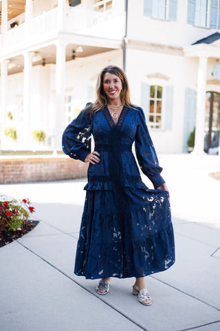 Woman smiling on a paved walkway in a navy blue floral burnout lace maxi dress featuring long sheer bishop sleeves, a scalloped lace V-neckline, a fitted waist with crochet lace trim, and a tiered ruffled maxi skirt. Styled with low block-heel metallic sandals, gold chain link necklaces, and a gold oval pendant.