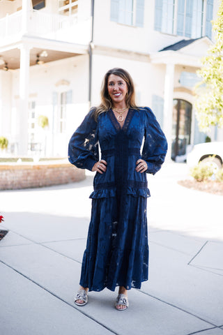Full-length front view of a woman posing with hands on hips in a midnight navy burnout lace long-sleeve maxi dress. Highlights the central crochet lace lattice trim running down the bodice, fitted waistline, layered ruffle accents across the hips, and tiered A-line skirt drape.