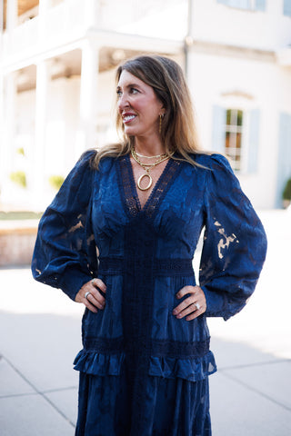 Mid-length front view showcasing the intricate floral burnout pattern and crochet lace trim on the navy blue maxi dress. Details include the sheer balloon sleeves with fitted cuffs, crocheted V-neck frame, and ruffled waist seam, complemented by chunky gold jewelry.