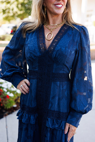 Upper body portrait highlighting the texture of the navy floral burnout jacquard fabric and scalloped lace neckline. Shows the detailed sheer sleeves, fitted waistline band, and layered gold chain statement necklace.