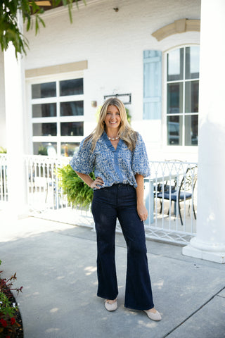 Woman posing outdoors in a light blue chambray-style blouse featuring intricate white embroidered geometric scrollwork patterns, a ruffle-trimmed V-neckline with a functional button placket, and voluminous elbow-length puff sleeves. Styled tucked into high-waisted dark wash flare denim jeans with neutral pointed-toe flats, a short beaded necklace, and gold hoop earrings in front of a white building background.