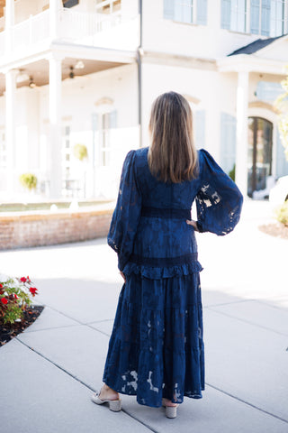 Full-length back view of the navy blue burnout lace maxi dress showing the tiered ruffled skirt construction, fitted waistline, and sheer volume of the long bishop sleeves from behind. Styled with brown hair worn loose and metallic low-heeled mules.