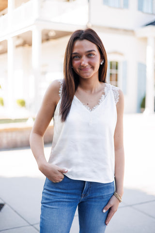 Woman smiling outdoors wearing a crisp white satin camisole top featuring intricate delicate eyelash lace trimming along the V-neckline and shoulder straps. Styled casually with mid-rise blue jeans and gold beaded wrist accessories set against a bright building backdrop.