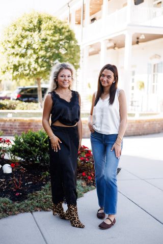 Two women posing together outdoors showcasing colorway styling options. One model wears the black eyelash lace satin cami with a matching black lace-trimmed satin skirt and leopard print boots; the other wears the white eyelash lace satin cami with classic blue jeans and brown Mary Jane flats