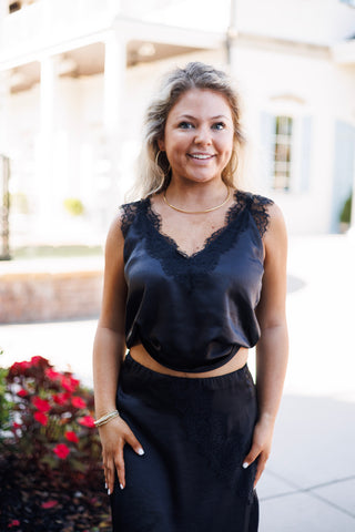 Woman smiling outdoors in a sleeveless black satin camisole top featuring delicate black eyelash lace trim along the V-neckline and shoulder straps. Styled with a matching high-waisted black satin midi skirt with lace side paneling and a sleek gold choker necklace.