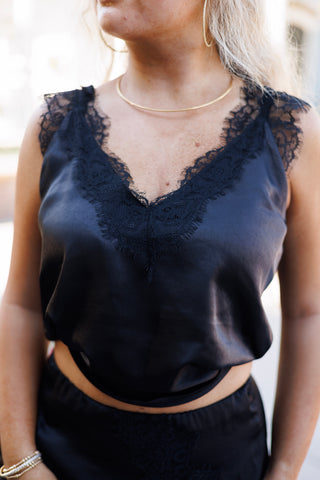 Upper body shot focusing on the chest detail of the black satin camisole top, showcasing the deep V-neckline accented by intricate eyelash lace fringe and wide lace-trimmed straps. Accessorized with gold hoop earrings and a minimal choker necklace.
