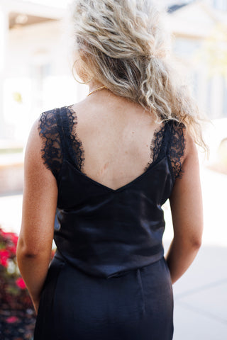 Back view from the waist up of the black satin cami top, displaying the deep V-back cut and thick eyelash lace strap detailing along the shoulders. Styled with blonde curly hair worn half-up.