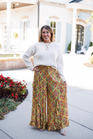 Woman smiling on a paved walkway in front of an upscale white building, posing with hand on hip in mustard yellow wide-leg palazzo pants featuring a vibrant pink and orange floral print, vertical cascading ruffles, a ruffled hemline, and a drawstring elastic waist. Styled with a cream ribbed crewneck sweater tucked in front, metallic open-toe sandals, and a gold link chain necklace with a circular pendant.