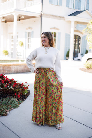 Full-length view of a woman showcasing the fluid movement and drape of mustard floral wide-leg pants with vertical ruffle trim and a ruffled hem. Paired with an off-white long-sleeve knit sweater, metallic slide sandals, and gold statement jewelry set against an elegant outdoor courtyard background.