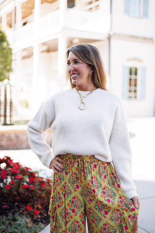 Three-quarter shot highlighting the details of the floral ruffle palazzo pants, including the elastic drawstring waist, lightweight printed fabric with red, pink, and orange blooms, and side ruffle accents. Styled with a cozy cream crewneck sweater and a gold link chain pendant necklace.