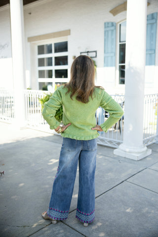 Back view of a woman wearing the long-sleeve lime green cardigan, showcasing the textured pointelle knit along the arms and the delicate ruffle trim at the hemline. Styled with hands on hips, highlighting the back pocket placement and embroidered bottom hem of the medium-wash wide-leg jeans on a sunny concrete patio walkway.