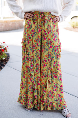 A detailed shot focusing on the high-waisted mustard yellow floral pants, highlighting the flexible drawstring elastic waistband, lightweight printed crepe fabric, vertical cascading ruffle trim along the legs, and the ruffled bottom hem. Paired with a cozy cream sweater and metallic low-heeled slide sandals.