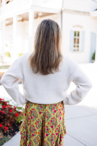 Back view from the waist up showing a cream ribbed crewneck knit sweater paired with high-waisted mustard yellow floral print wide-leg pants featuring a gentle ruffle along the upper hip and waistline.