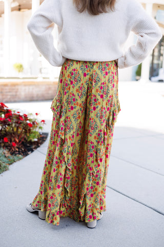 Back view from the shoulders down showcasing the dramatic wide-leg silhouette and full drape of the mustard floral pants, detailing the continuation of the vibrant pink floral pattern and cascading side ruffles down to the floor-length ruffled hem. Styled with hands on hips wearing a cream ribbed long-sleeve knit top.