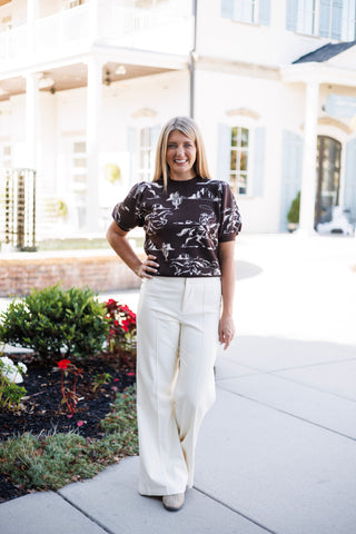 Woman posing outdoors in a dark brown short-sleeve knit sweater featuring a white Western print with cowboys, horses, and cacti. The puff-sleeve top is tucked into high-waisted, wide-leg cream trousers with center seam details, styled with beige closed-toe shoes against a bright courtyard background.