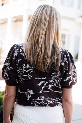 Back view of a woman wearing a dark brown short-sleeve knit sweater showcasing the continuous white Western toile print across the back and gathered puff sleeves. Styled with high-waisted cream wide-leg trousers.