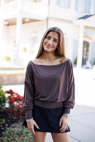 Woman smiling outdoors wearing a dark espresso brown long-sleeve knit top featuring a wide off-the-shoulder boat neckline, relaxed dolman sleeves, gathered cuffs, and a wide banded hemline. Styled with a black mini skirt, delicate cross necklace, stackable bracelets, and a silver watch against a bright courtyard backdrop