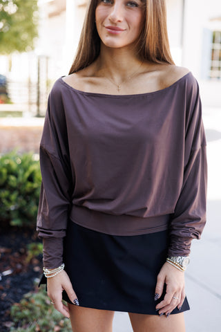 A portrait view of the off-the-shoulder dolman top highlighting the graceful wide boat neck that sits gently off the collarbones. The ultra-soft jersey knit fabric drapes effortlessly into a blouson silhouette over a fitted bottom band, complemented by a thin gold cross pendant and layered beaded bracelets