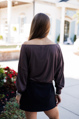 Back view of the espresso brown long-sleeve top showcasing the wide neckline cut across the upper back and shoulders. The slouched, fluid drape of the dolman sleeves gathers neatly at the wide hemline, paired with a black tailored mini skirt and straight brown hair worn loose.