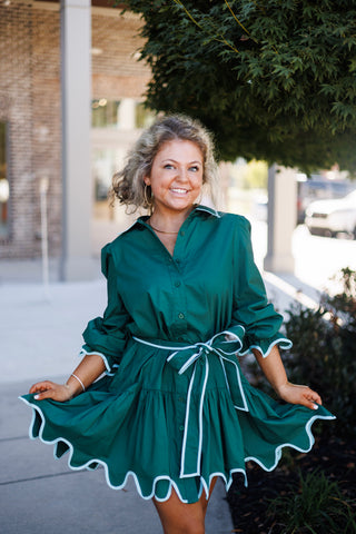 Three-quarter portrait of a smiling woman holding out the flared skirt of the Emerald Scalloped Shirt Dress. The rich green poplin dress showcases a pointed collar, button-down bodice, push-up three-quarter sleeves with contrast-piped ruffle trim, and a tiered A-line mini skirt.