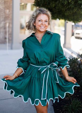 Front-facing mid-shot highlighting the structured A-line silhouette and white/light blue contrast edge trim on the Emerald Scalloped Shirt Dress. The model holds the scalloped hem wide, emphasizing the volume of the tiered skirt and the tied waist belt.