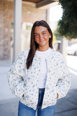 Woman smiling outdoors in a cozy, cream and light blue floral print quilted bomber jacket featuring a relaxed silhouette, drop shoulders, and a collarless neckline. Worn open over a solid white crewneck top and medium-wash denim jeans with hands resting in the side pockets against a paved walkway backdrop.