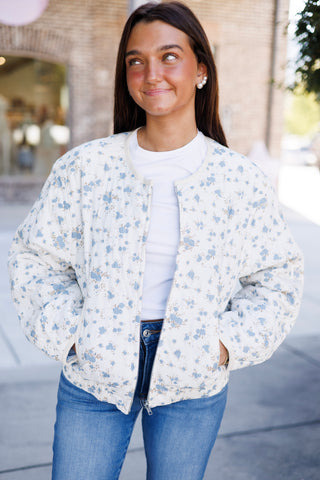 Three-quarter portrait view of a woman wearing a white floral quilted zip-up jacket adorned with delicate dusty-blue floral sprigs. The casual jacket features a cream trim collarless neckline, silver front zipper hardware, and elasticized ribbed trim along the hem, paired with medium-wash blue jeans.