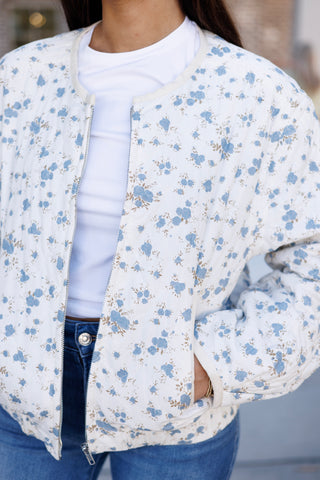 Close-up shot highlighting the diamond quilting, soft cream trim, and silver front zipper closure of a white floral printed bomber jacket. Details showcase the subtle blue floral pattern texture, side welt pockets, and layering over a classic white t-shirt and blue denim waistband.   