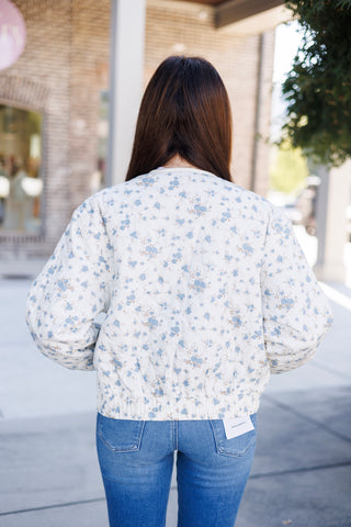 Back view of a woman wearing a white and light blue floral quilted zip jacket showing the uninterrupted floral diamond-quilted pattern across the back and slouchy sleeves. The jacket is styled with an elasticized gather hem over blue denim jeans.   