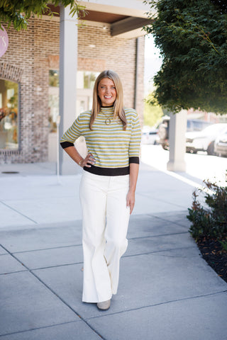 Woman smiling and standing on a sidewalk in front of a brick storefront, posing with hand on hip in a multi-colored striped fine-knit top featuring horizontal olive green, powder blue, white, and heather grey stripes. The top features contrast dark brown ribbed trim along the mock neckline, elbow-length cuffs, and hem, paired with high-waisted off-white wide-leg trousers, a chunky gold ring pendant necklace, and grey pointed-toe boots.