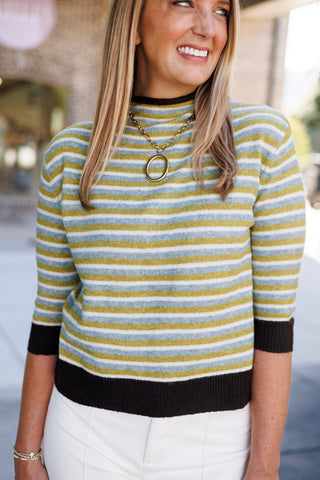 Close-up portrait view of the fine-gauge striped knit top showing the soft texture and subtle horizontal pattern of olive green, powder blue, and white stripes. Accentuated by a dark brown ribbed mock collar, three-quarter length sleeves with matching ribbed cuffs, and a bold gold oval hoop necklace.