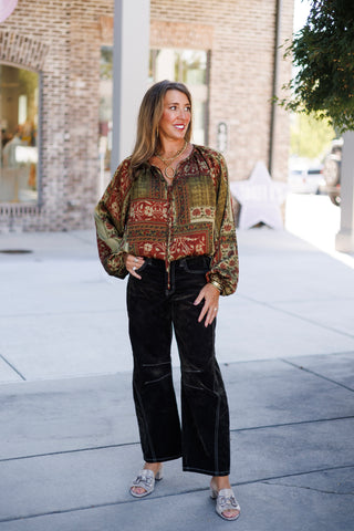 Woman standing outdoors on a sidewalk wearing a relaxed-fit bohemian patchwork blouse featuring intricate rust red, olive green, and beige floral tapestry prints with long balloon sleeves and a split neckline with ties. The blouse is tucked into dark espresso brown wide-leg corduroy pants with white contrast stitching, accessorized with layered gold chain necklaces, a ring pendant, and metallic mule sandals.