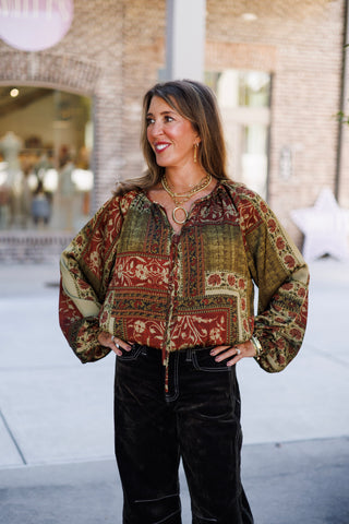 Medium full-length shot of a woman posing with hands on hips wearing a bohemian paisley and floral patchwork blouse in rich earth tones of rust red, olive green, and tan. Features gathered volumetric billowy sleeves, a split v-neckline with neck ties, and dark velvet corduroy wide-leg pants with detailed contrast seam stitching.