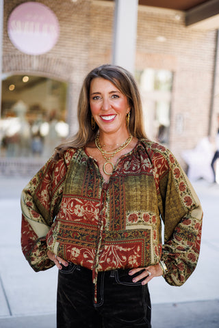 Close-up shot highlighting the texture and detail of a printed boho peasant blouse featuring a split keyhole neckline with ties, full balloon sleeves, and an eclectic combination of rust red and olive green floral tapestry patterns. Accessorized with gold drop earrings and a chunky gold link chain necklace.