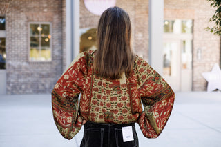 Back view of the bohemian printed blouse from the waist up, displaying the continuous rust red and olive green floral patchwork design across the back and voluminous long sleeves. Paired with high-waisted dark brown corduroy pants featuring white contrast stitching and a back pocket brand tag.   