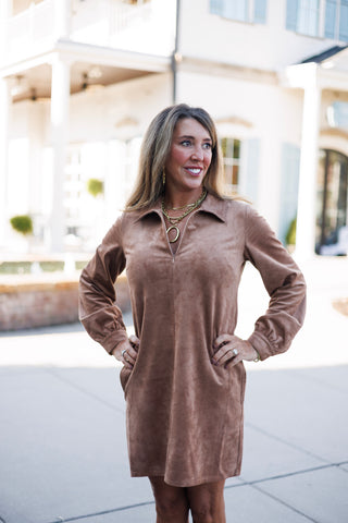 Front-facing mid-shot highlighting the spread lapel collar and split V-neckline of a taupe brown faux-suede mini dress. Posed with hands on hips, showcasing the gathered sleeve cuffs, layered gold link necklaces with a large oval charm, and gold statement earrings.